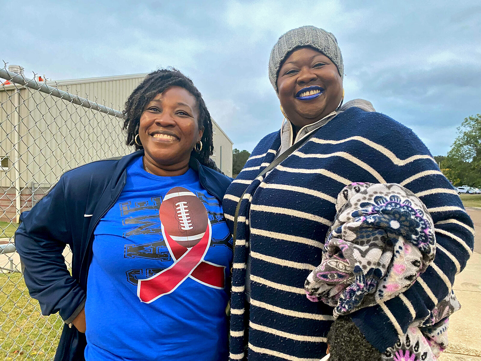 Murrah High School student athlete parents Lawanda Gholar and Erin Phillips stand for a picture together before their sons' game against Madison Central High in Madison, Mississippi. Gholar is on the left and is wearing a blue Murrah t-shirt, while Phillips is on the right wearing a blue and white sweater and blue lipstick. Both women are happy their school institute a COVID vaccine mandate for student athletes.