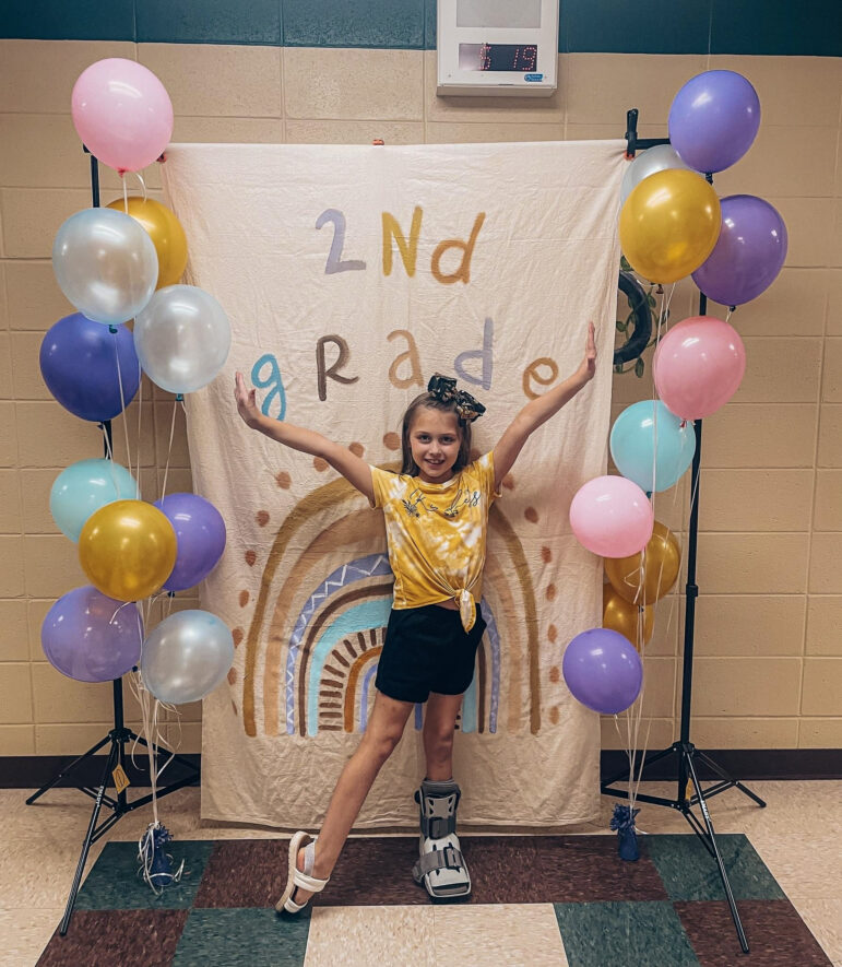 Layla Hill, 8, stands in front of a sign that says "2nd Grade" while wearing a walking boot.